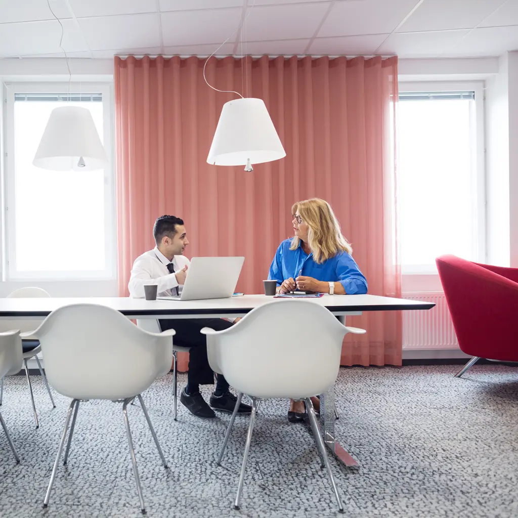 Personer sitter vid bord i ljus kontorsmiljö med rosa gardiner och modern inredning.